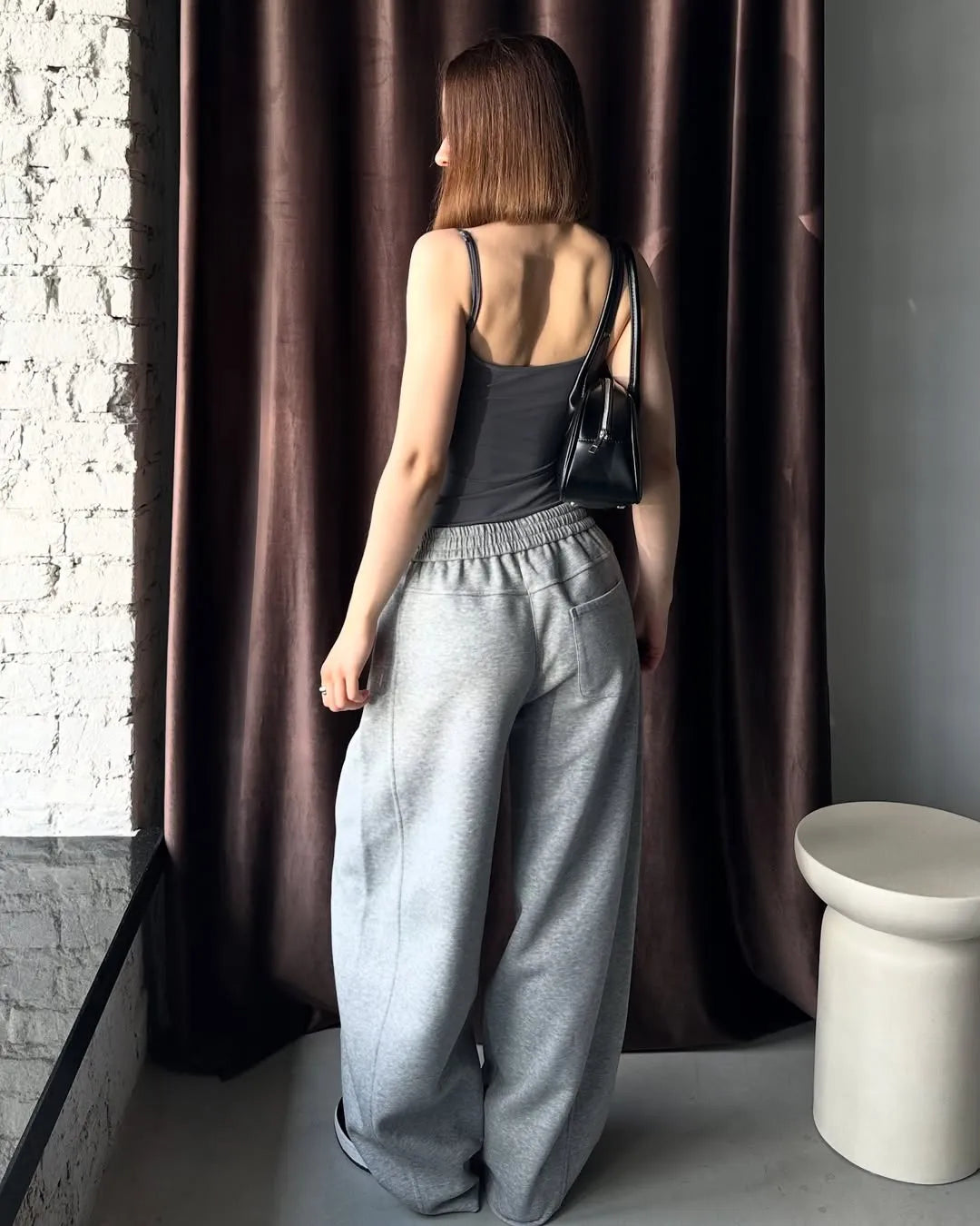 Woman in grey sweatpants standing against a brown curtain.