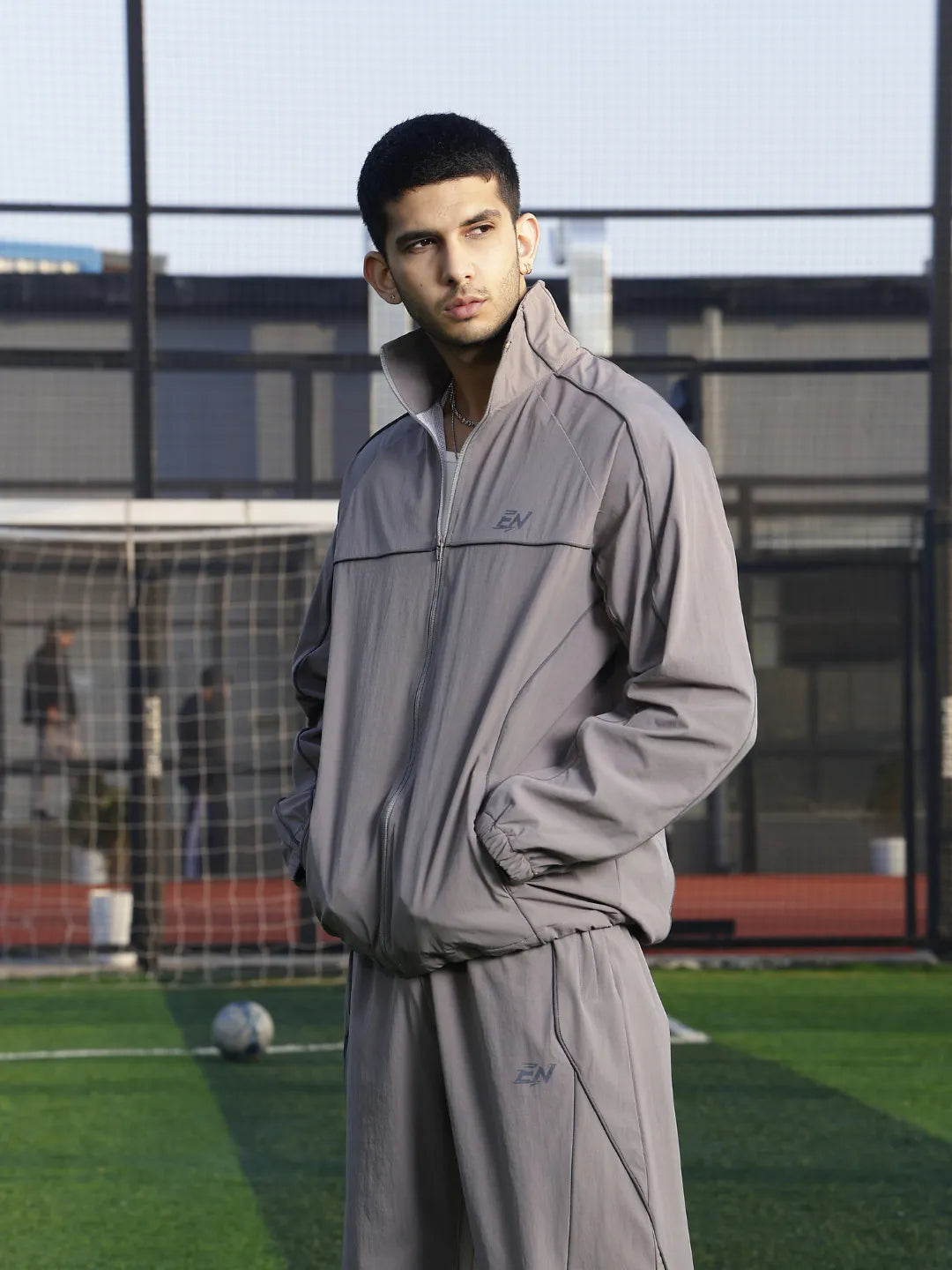 Person wearing a grey sports jacket  on a sports field with a soccer ball and goalpost in the background.