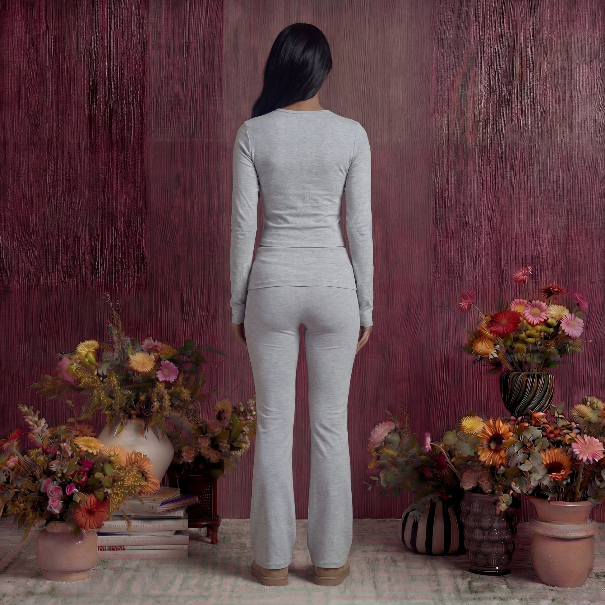Woman in a grey cotton fold over pants and top standing in front of a textured purple wall with floral arrangements.