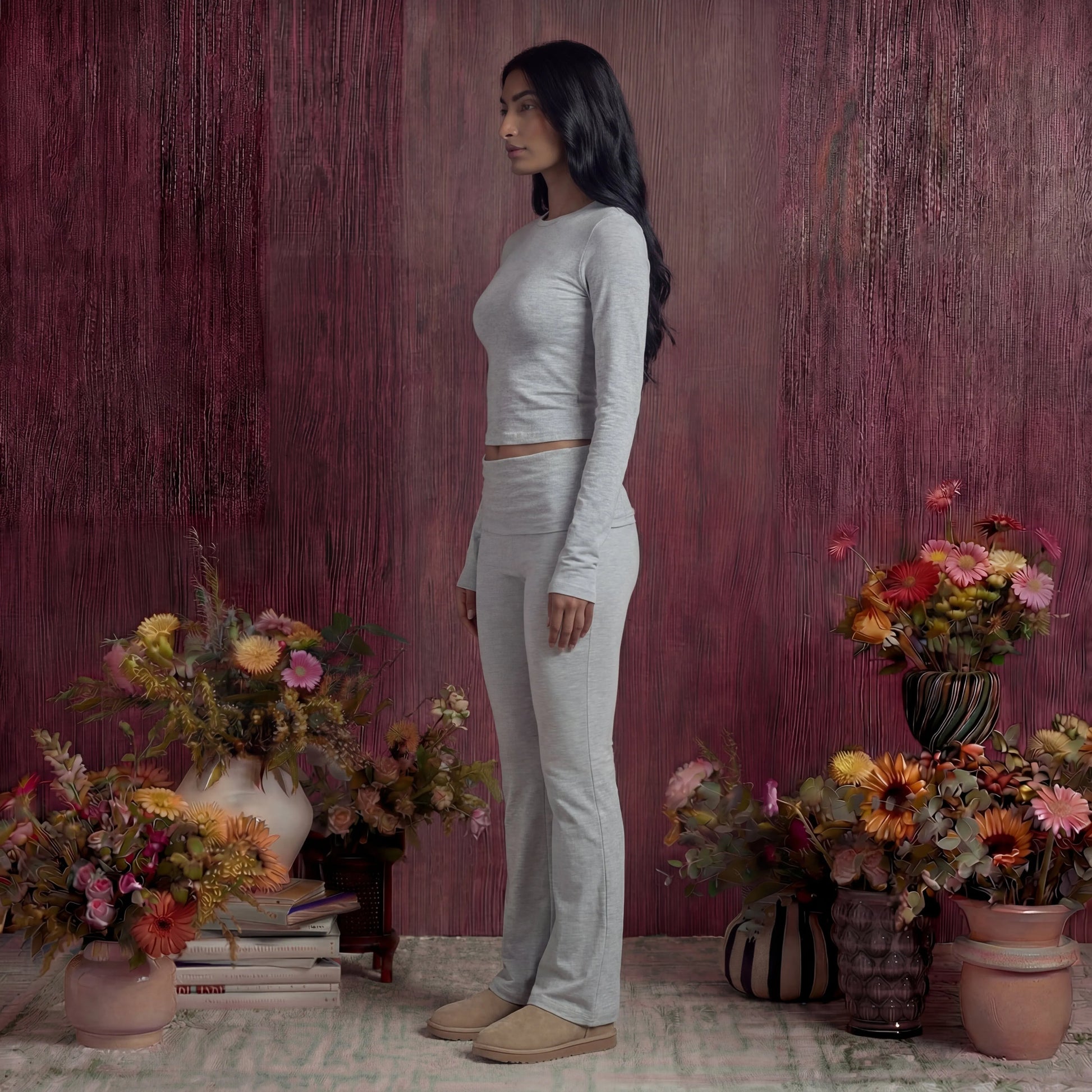 Woman in a light grey cotton fold over pants and top standing among potted flowers against a textured purple wall.