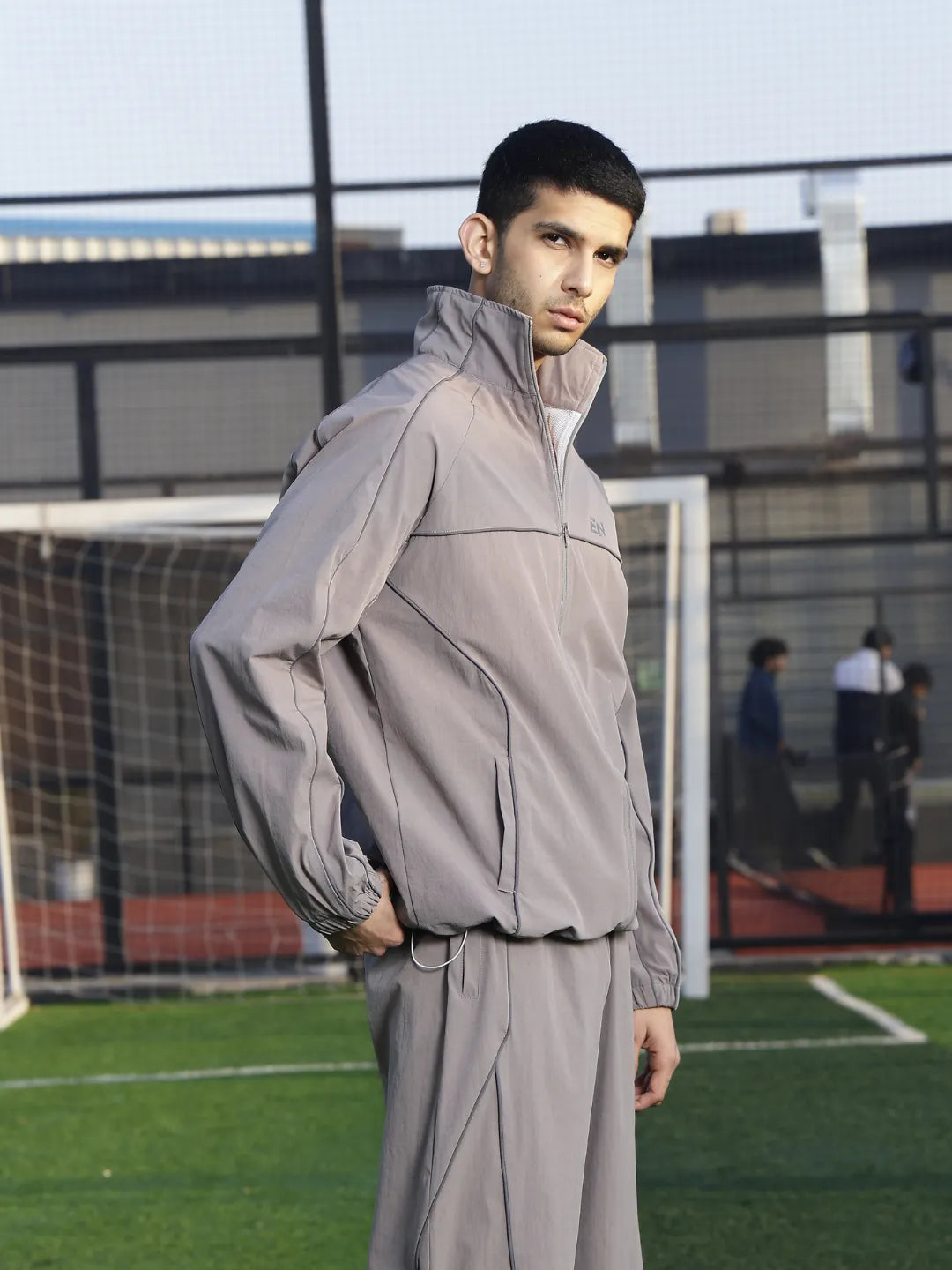 Person wearing a grey sports jacket  on a sports field with a goalpost in the background