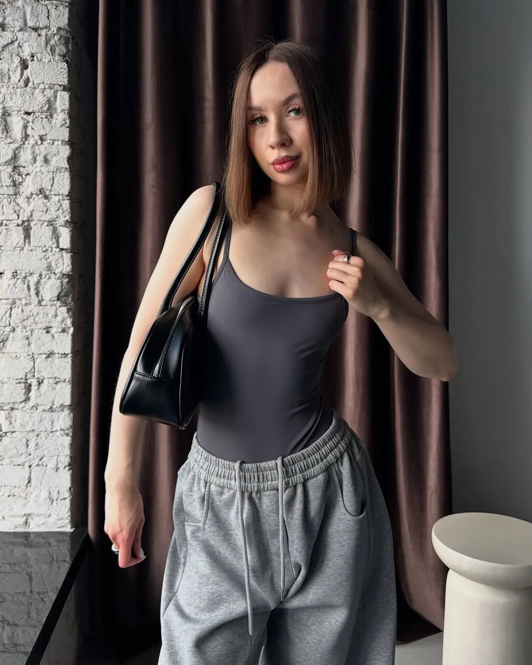 Woman in grey sweatpants standing against a brown curtain.