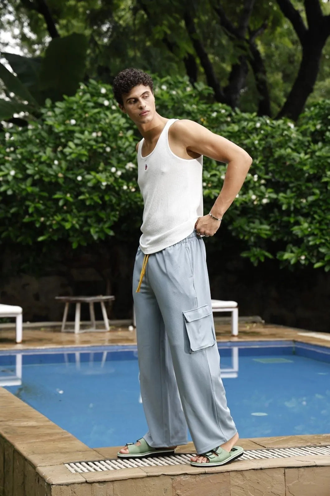 Man in a white tank top and light grey pants standing by a pool with greenery in the background