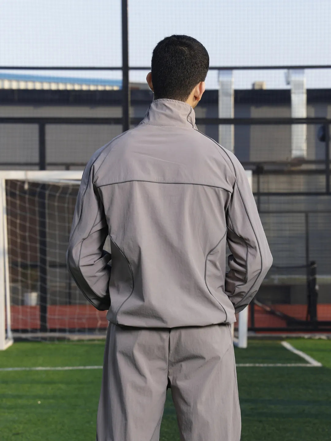 Person wearing a grey sports jacket  standing on a soccer field with a goalpost in the background.