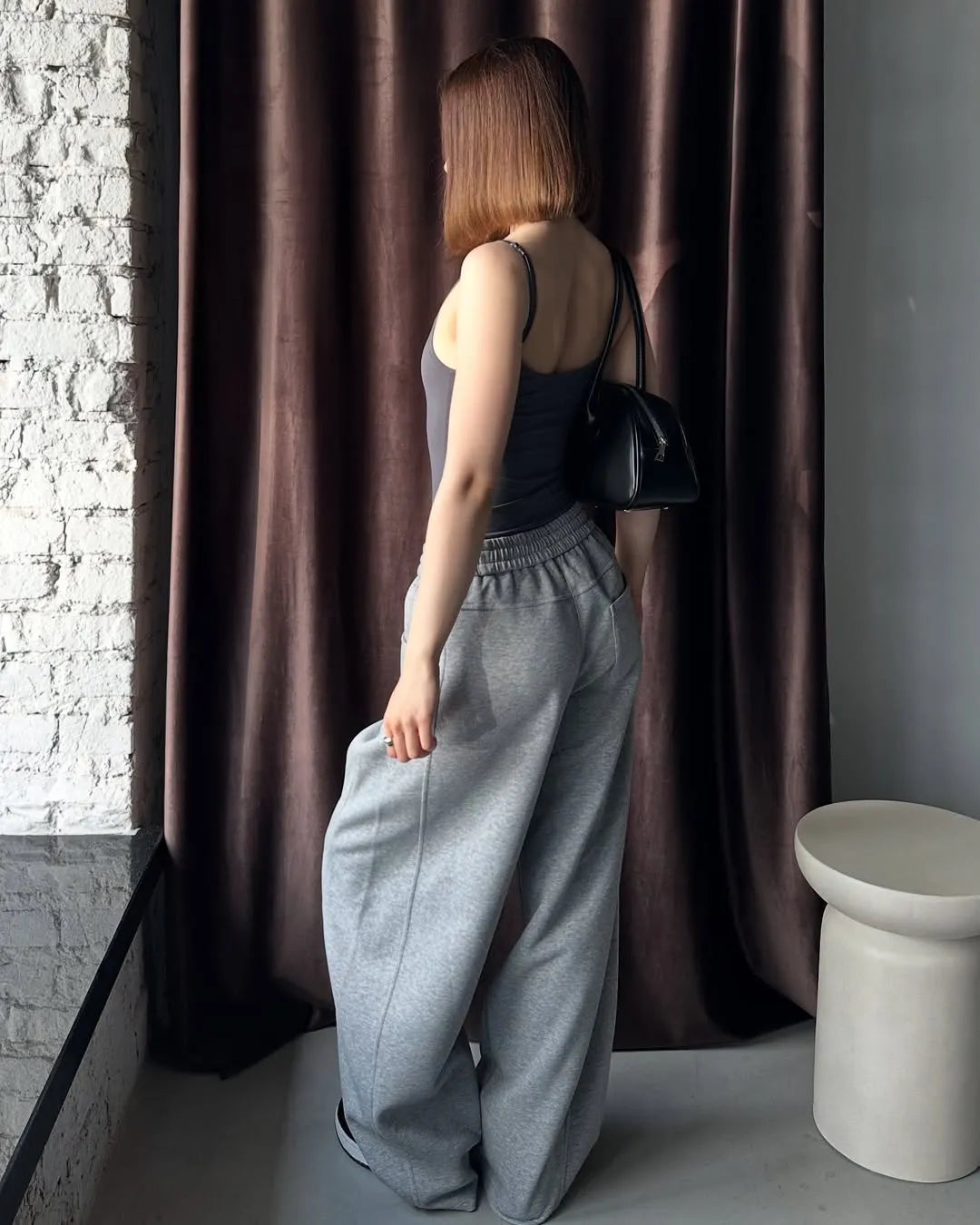 Woman in grey sweatpants standing against a brown curtain.