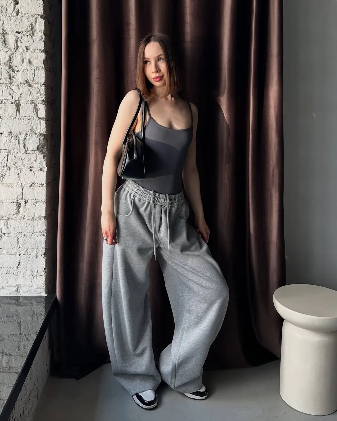 Woman in grey sweatpants standing against a brown curtain.