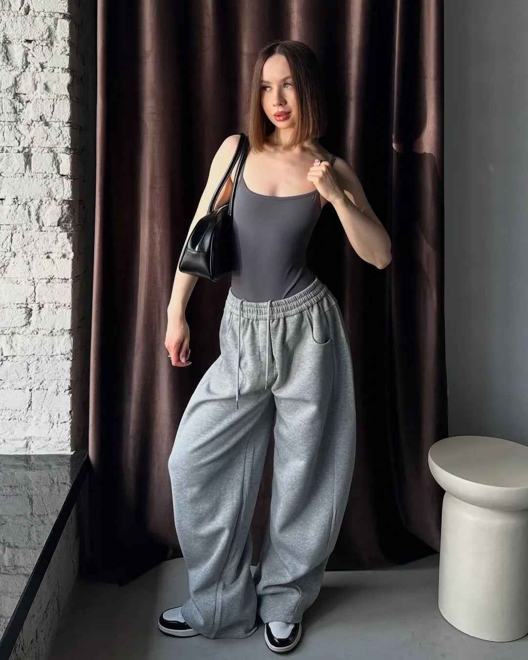 Woman in grey sweatpants standing against a brown curtain.