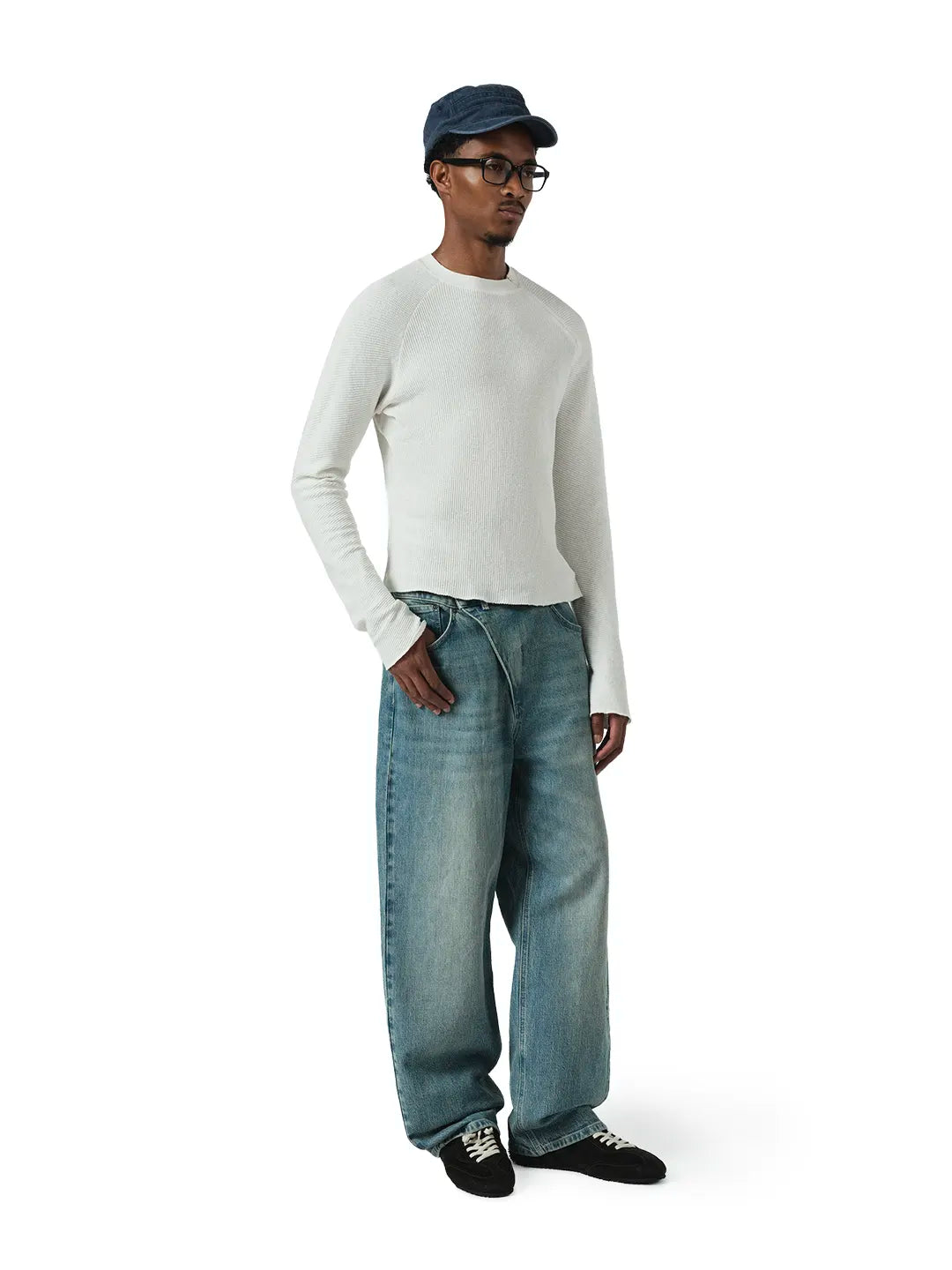 A model wearing a white long-sleeve knit sweater, wide-leg blue jeans, black sneakers, and a navy blue cap, standing against a plain white background.