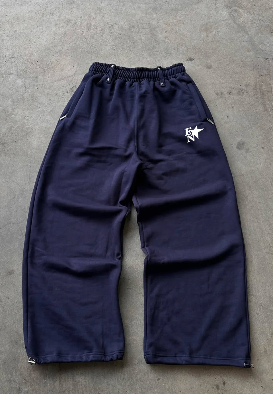 A pair of dark navy blue sweatpants with an elastic waistband and side pockets, laid flat on a grey concrete surface. A white star logo with the letters 'ER' is printed on the left hip.