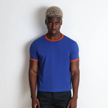 A man wearing a royal blue ringer t-shirt with orange trim on the crew neck and sleeve cuffs, paired with dark jeans, standing against a plain white wall.