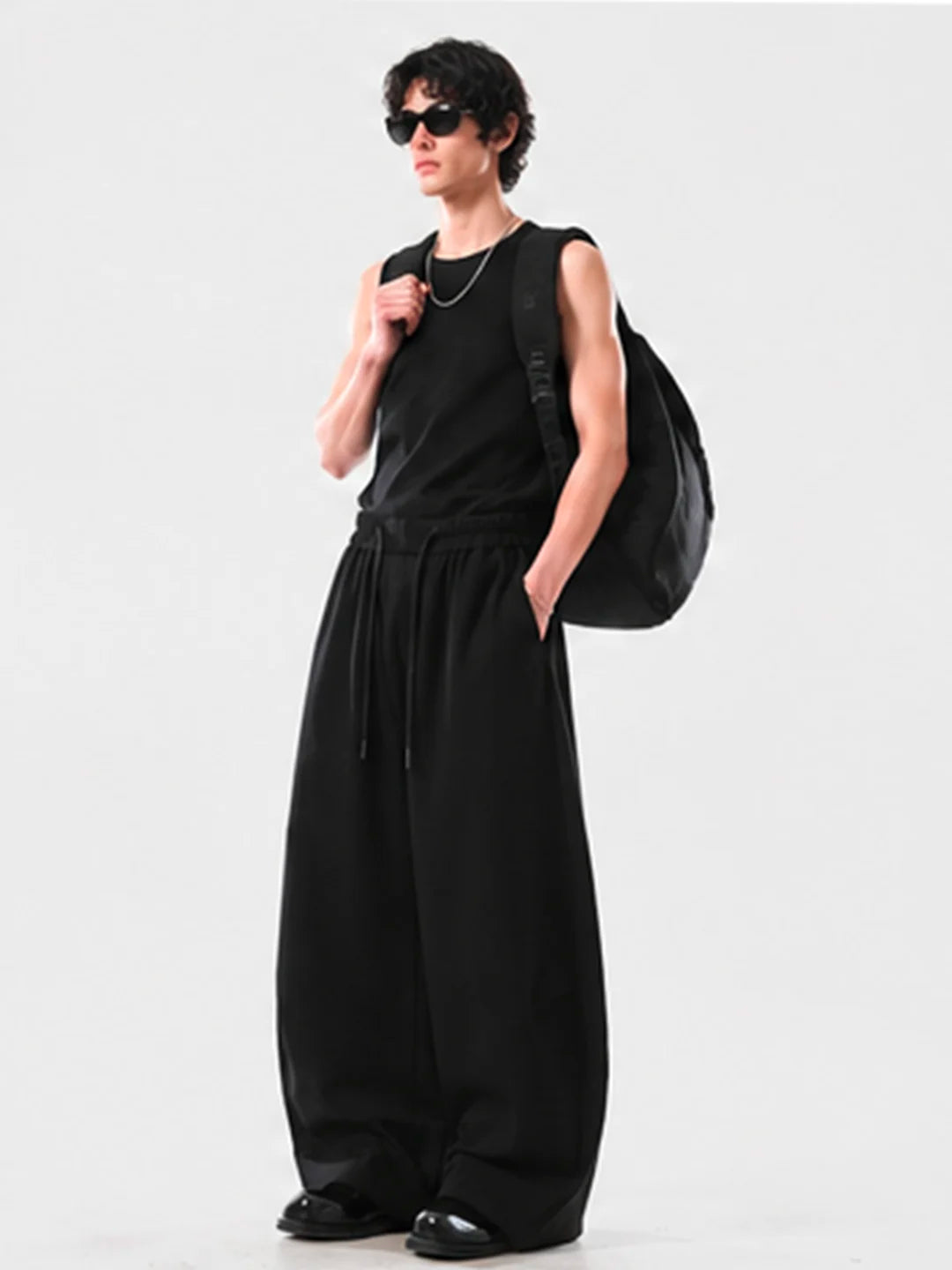 Person wearing a black sleeveless top and long black sweatpants, holding a black bag on a white background