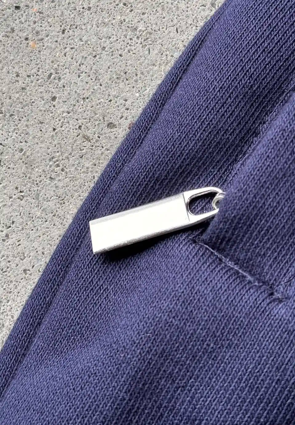 A silver metal zipper pull with a small integrated keyring, shown on a close-up of a navy blue textured fabric next to a grey concrete surface.