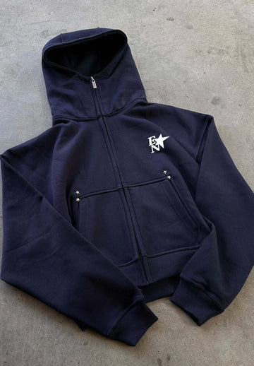 A navy blue zip-up hoodie with a white embroidered logo on the left chest, laid flat on a grey concrete surface.