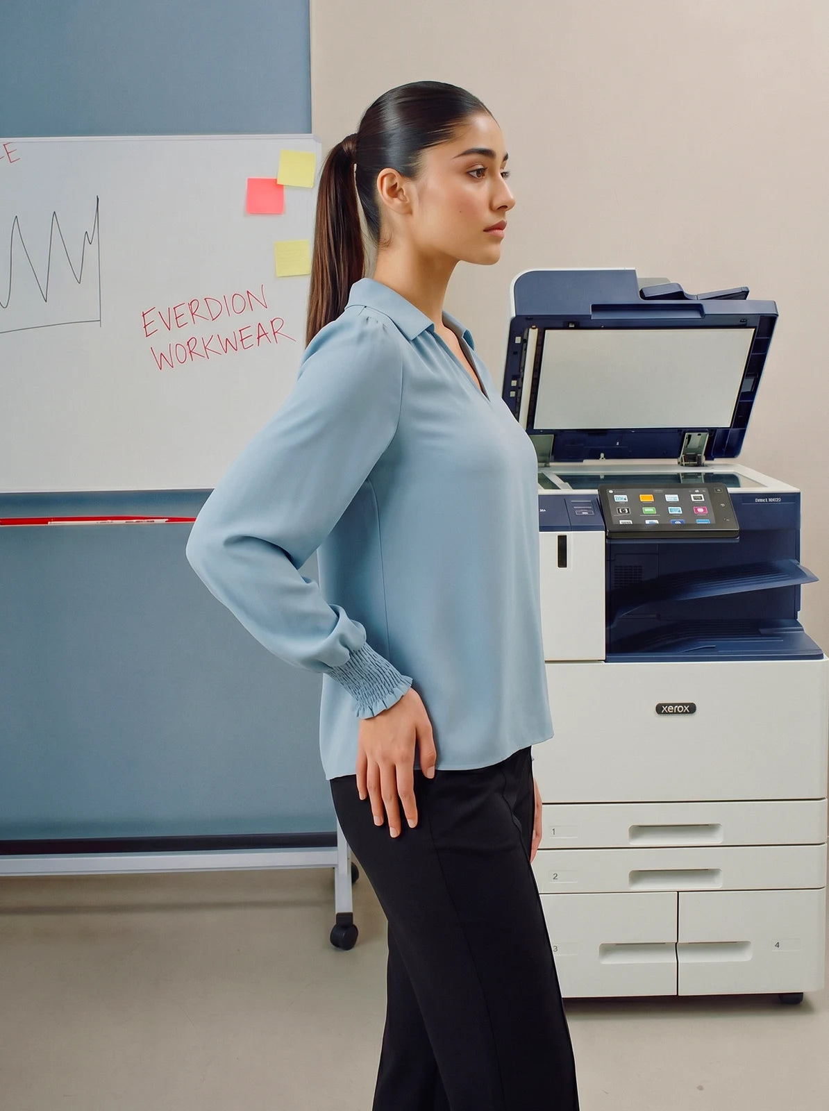 Person wearing a light blue blouse standing in an office setting with a printer and whiteboard in the background.