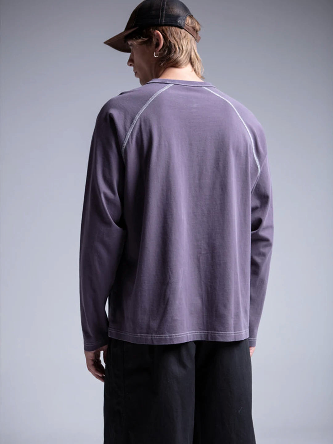 A person wearing a long-sleeve crewneck t-shirt in a lavender color, paired with black pants and a dark baseball cap