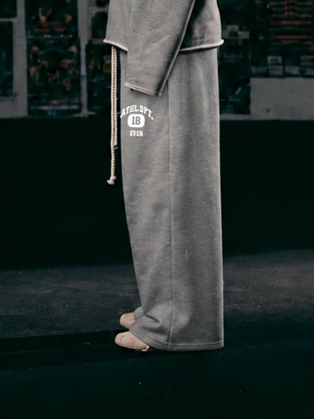 A person wearing oversized grey sweatpants with a white printed logo reading 'MELTY 18 ESTX' and a drawstring, shown from the waist down in a dark indoor setting.