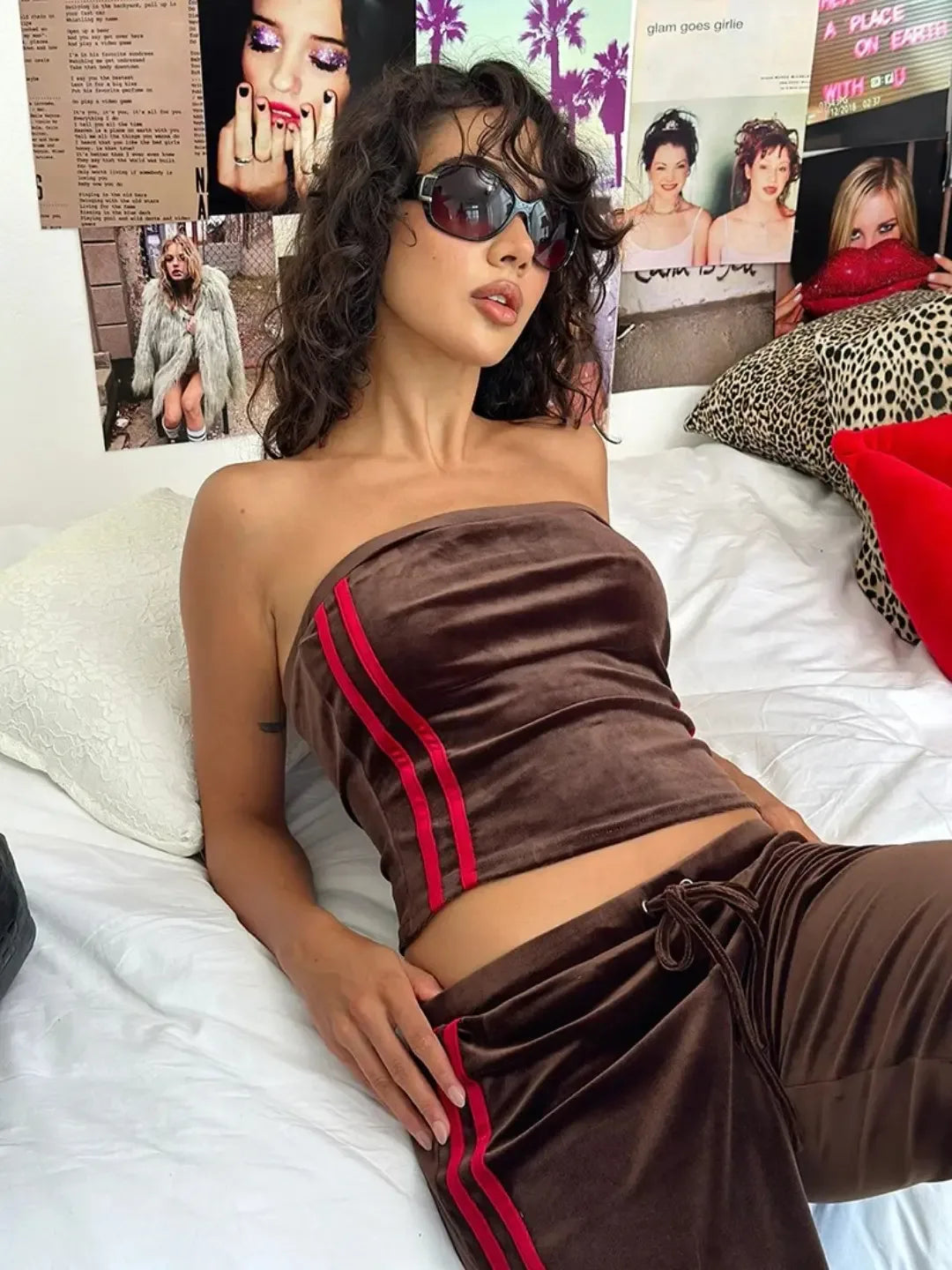 Woman in a brown outfit with red stripes sitting on a bed with magazine covers in the background.
