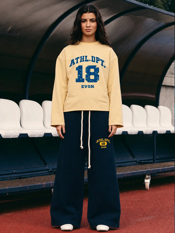 A woman wearing a cream tshirt with blue varsity lettering that reads 'ATHLDEPT. 18 EVON' and navy wide-leg sweatpants with a yellow and blue 'ATHLDEPT.