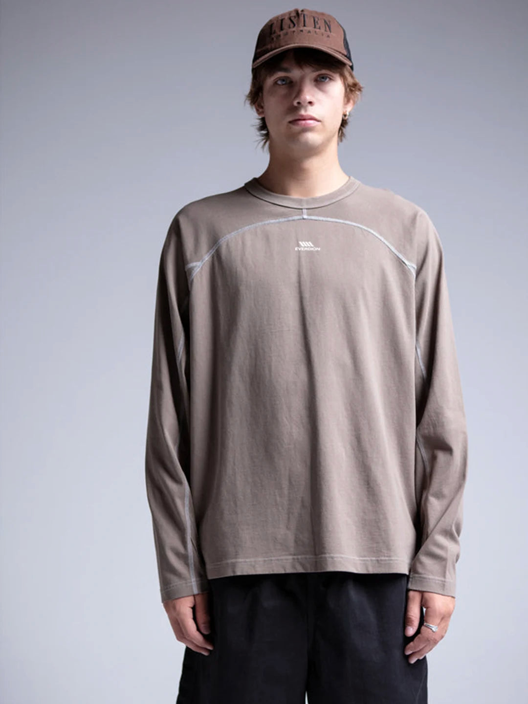 A model wearing a taupe brown long-sleeve crewneck shirt with white contrast stitching and a small white logo on the chest, paired with black pants and a brown cap.