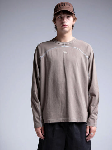 A model wearing a taupe brown long-sleeve crewneck shirt with white contrast stitching and a small white logo on the chest, paired with black pants and a brown cap.