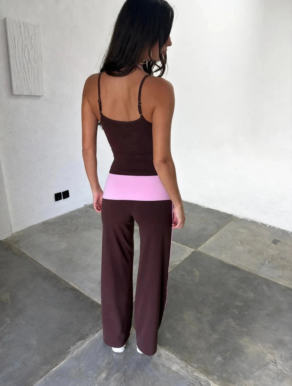 A woman wearing a dark brown two-piece activewear set with a wide light pink waistband, standing in a minimalist room with concrete floors and white walls.