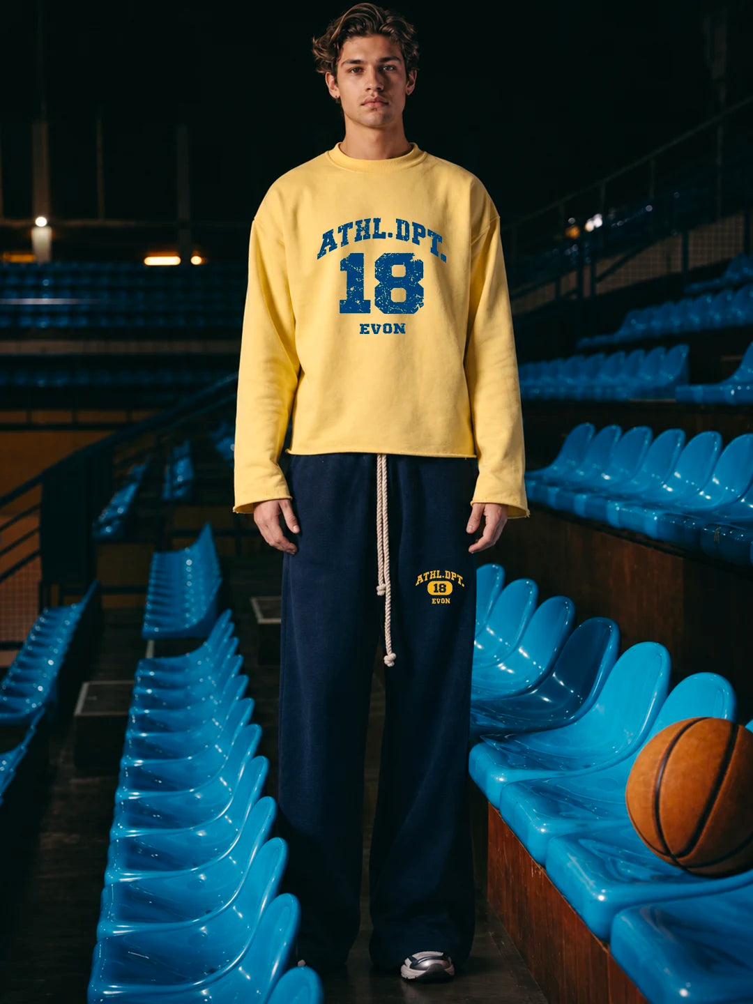 A man wearing a yellow long-sleeve sweatshirt with blue collegiate-style lettering that reads 'ATHL. DPT 18 EVON' and dark navy sweatpants with a drawstring and a small yellow logo.