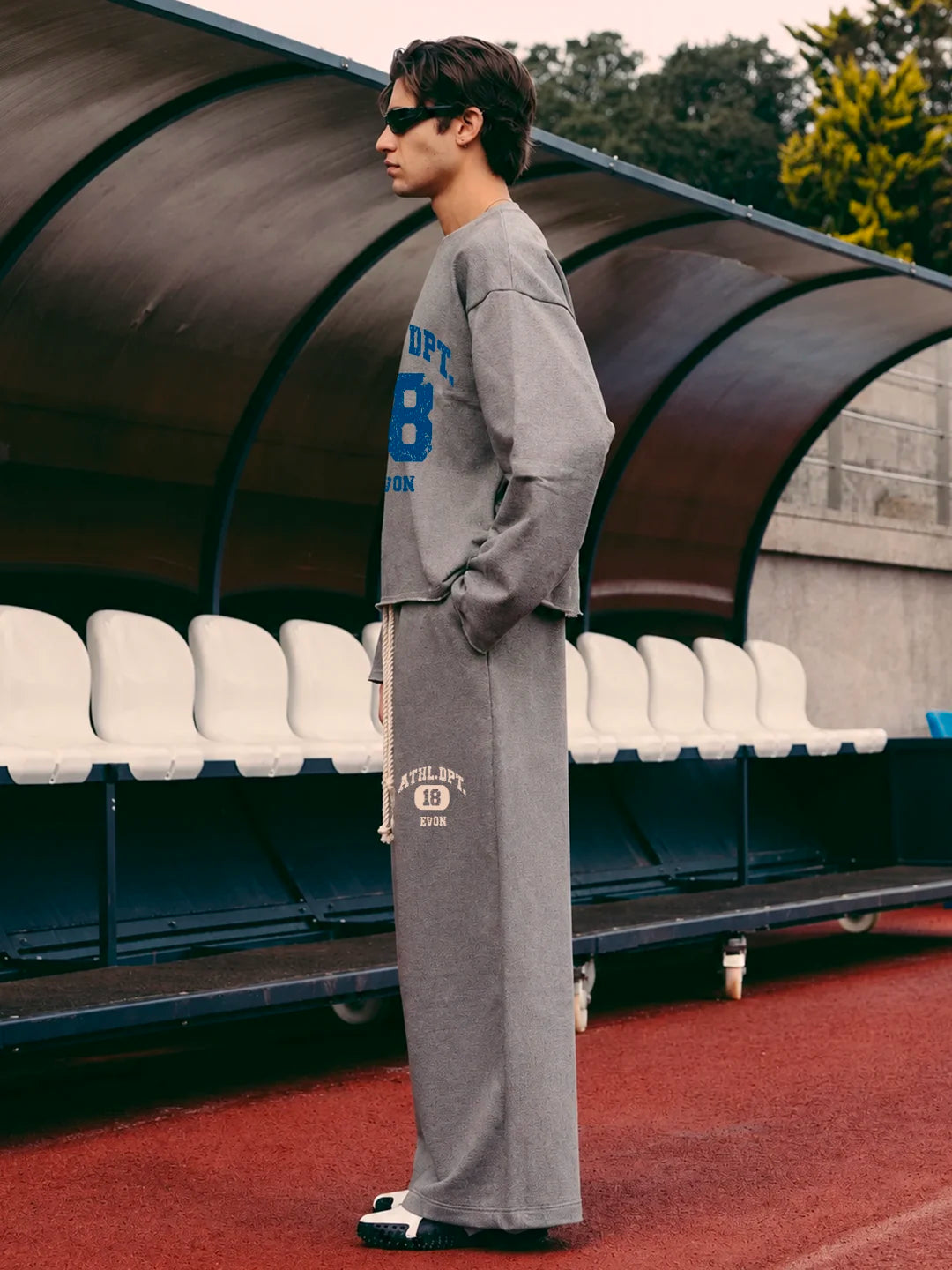 A man wearing a grey long-sleeve tshirt and matching sweatpants stands in profile in front of a stadium seating structure. The sweatshirt has blue and white lettering, and the sweatpants have white lettering near the lower leg.