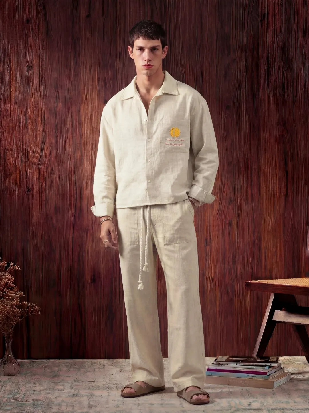 Man wearing a light-colored shirt and pants standing against a wooden wall.