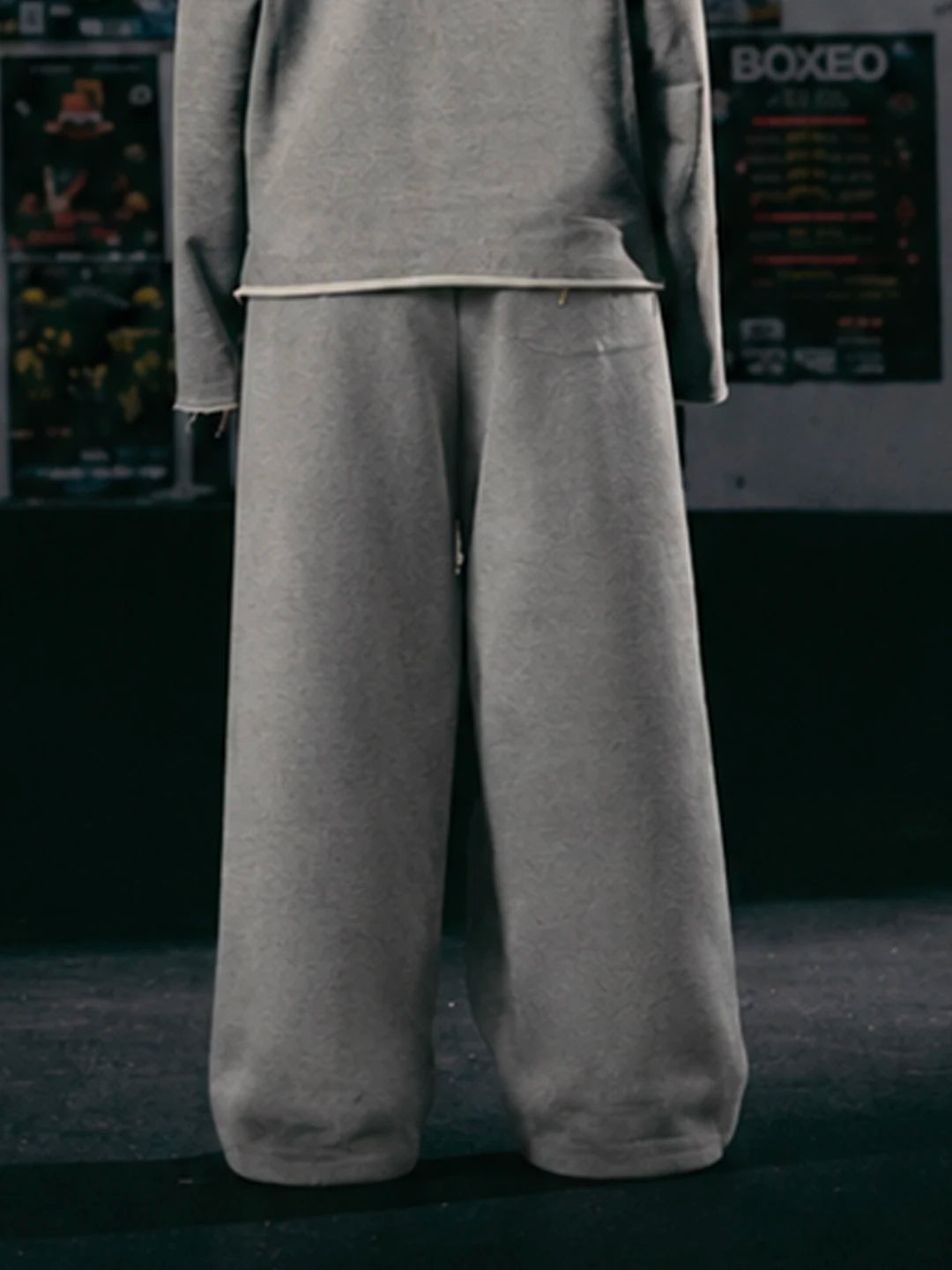 A person wearing a matching grey sweatshirt and very wide-leg sweatpants, shown from the back in a dark indoor setting with posters on the wall.