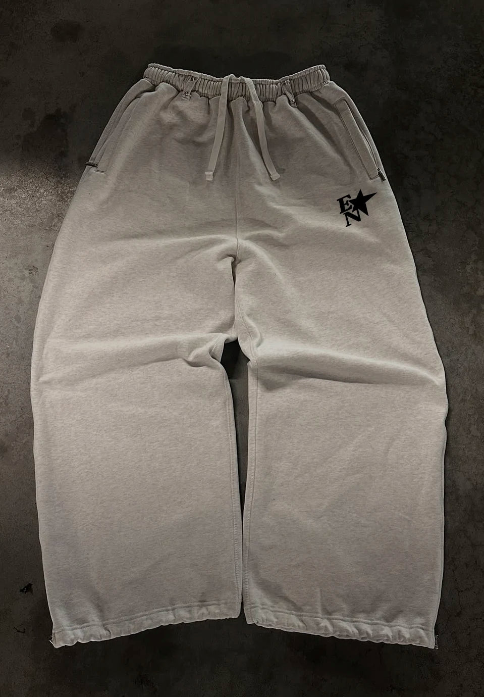 Light grey sweatpants with a drawstring waist and a small black graphic logo on the left hip, laid flat on a dark textured surface.