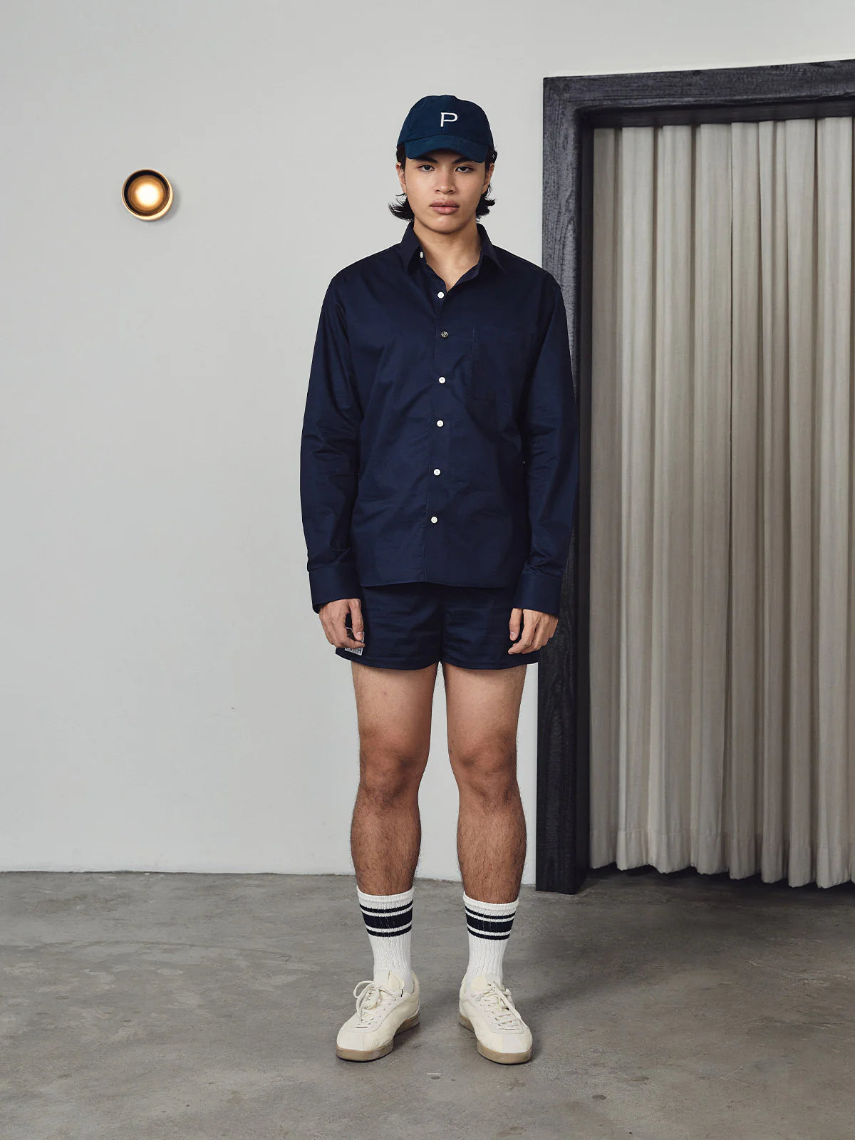 Person wearing a navy outfit with a cap, shirt, shorts, and white shoes in a minimalistic setting.