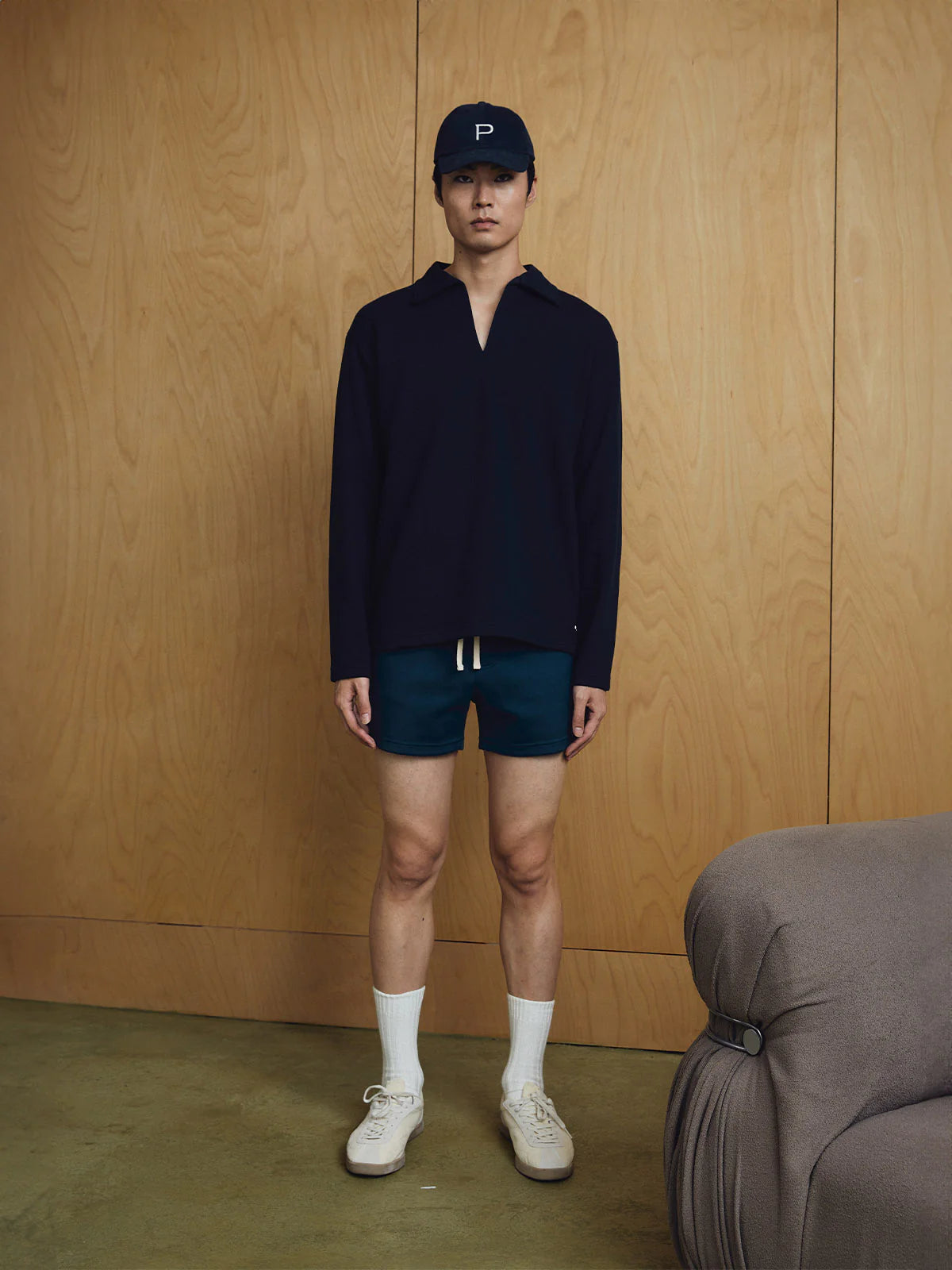 Person wearing a dark long-sleeve shirt, shorts, and a cap against a wooden wall.