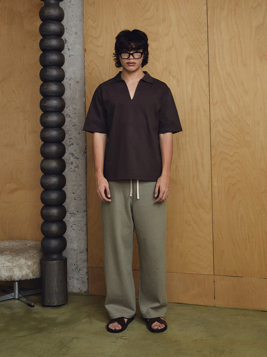 Person wearing a dark brown shirt and light grey pants standing against a wooden wall.