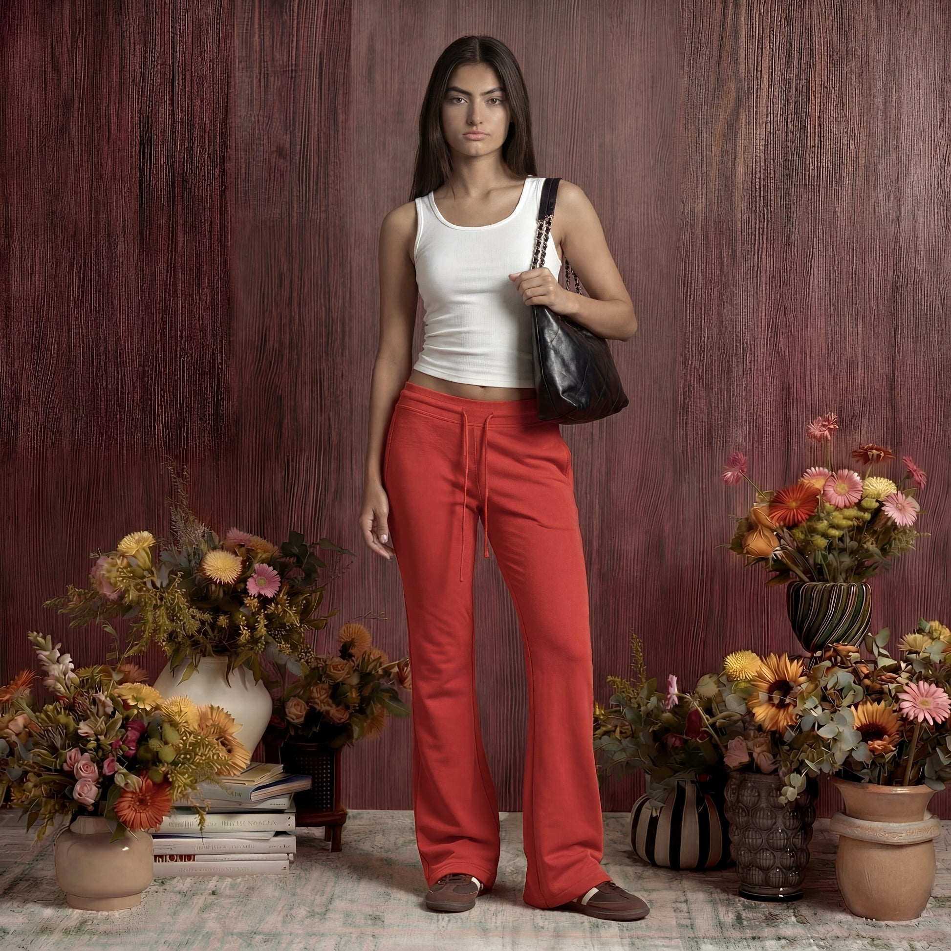 Woman in white top and red sweatpants standing among floral arrangements against a wooden wall.
