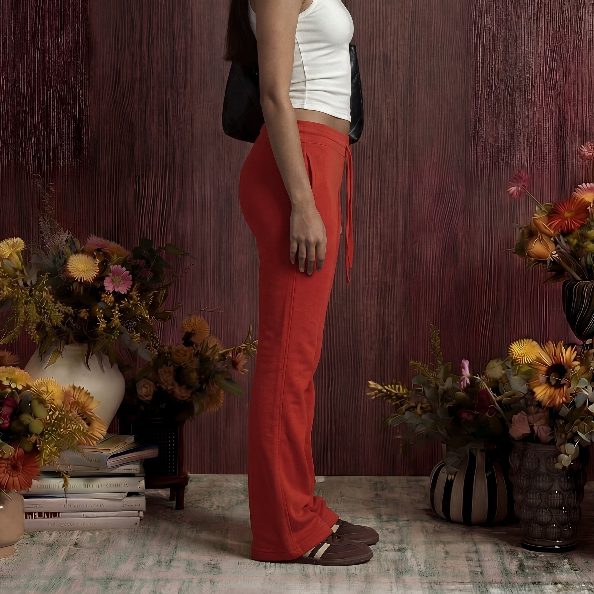 Person wearing red sweatpants standing among floral arrangements and books against a wooden wall.