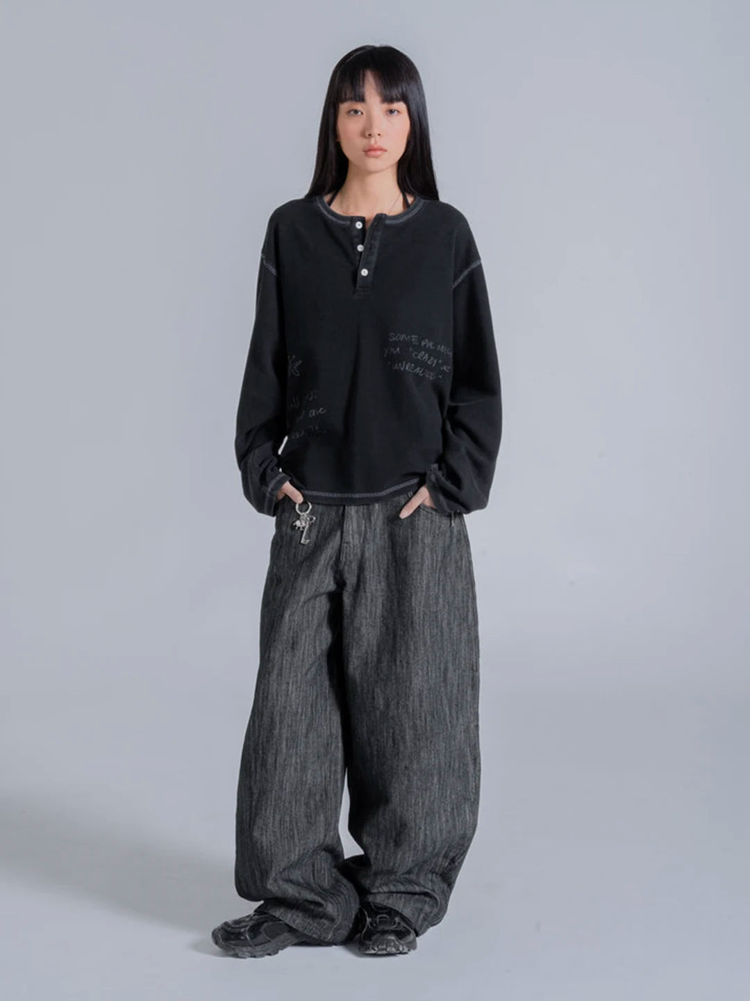 A model wearing a black cropped henley tshirt with tonal text graphics and very wide-leg dark grey textured pants.