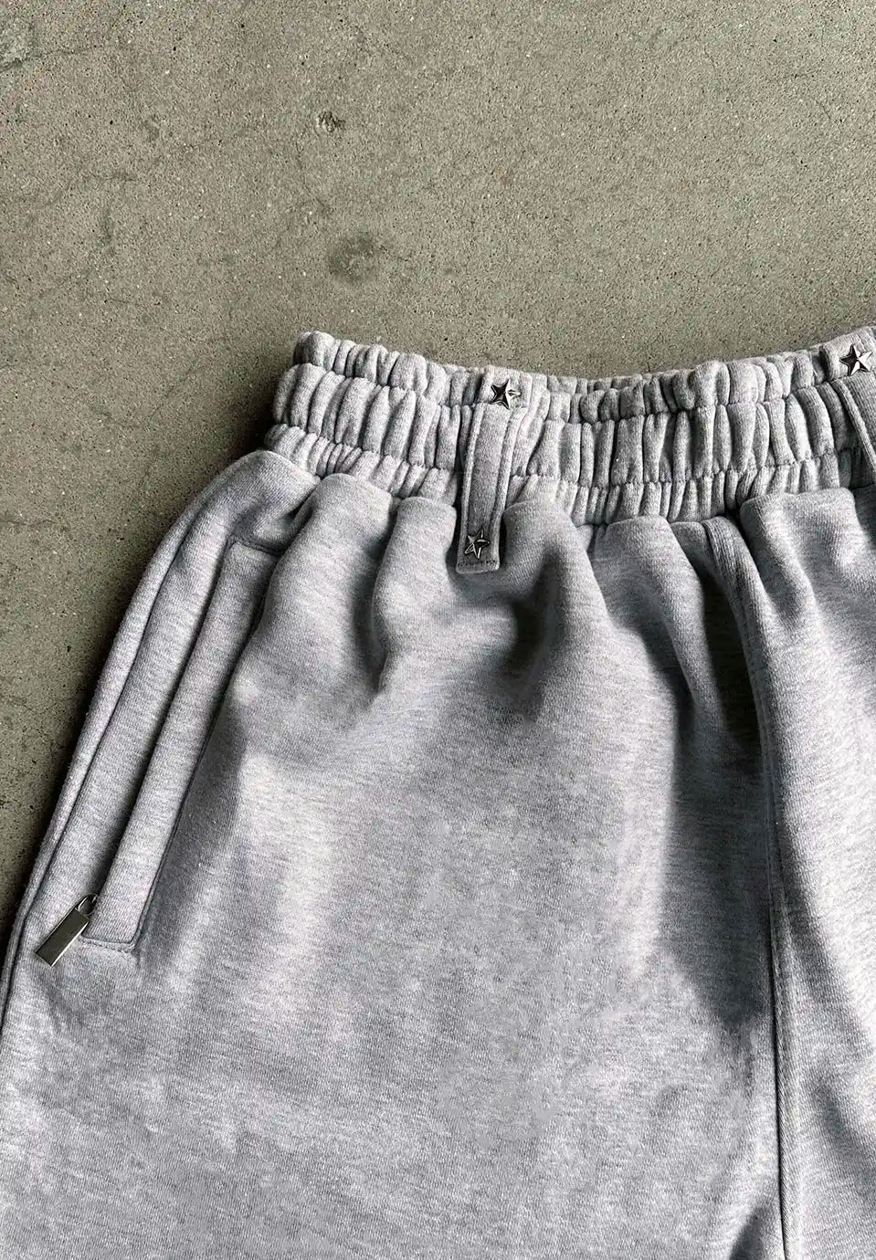 Close-up of light grey heather sweatpants laid flat on a textured concrete surface, showing the elastic waistband, drawstring detail, and side pocket.
