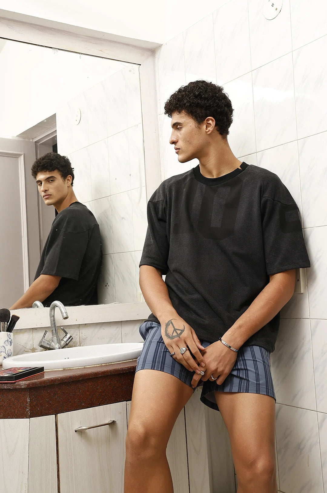 Person wearing a black t-shirt and plaid shorts standing in a bathroom.
