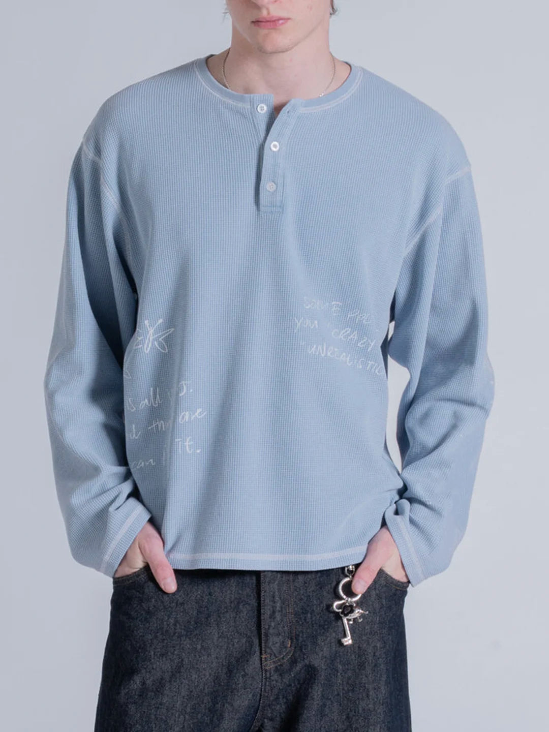 A person wearing a light blue ribbed long-sleeve henley t-shirt with white handwritten-style text and floral doodles, paired with dark jeans 