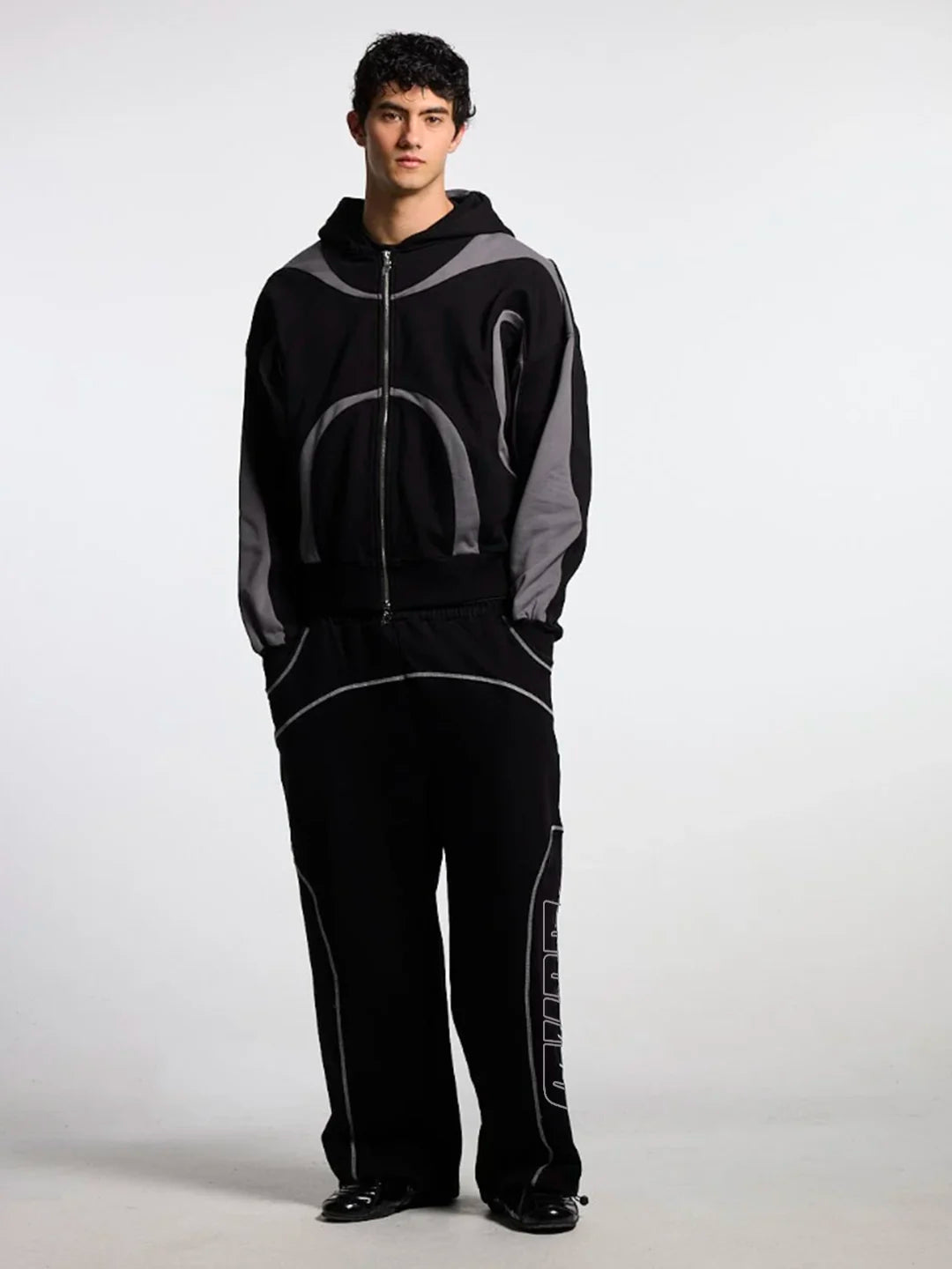 A man wearing a black and grey zip-up hoodie and matching black sweatpants with white piping details