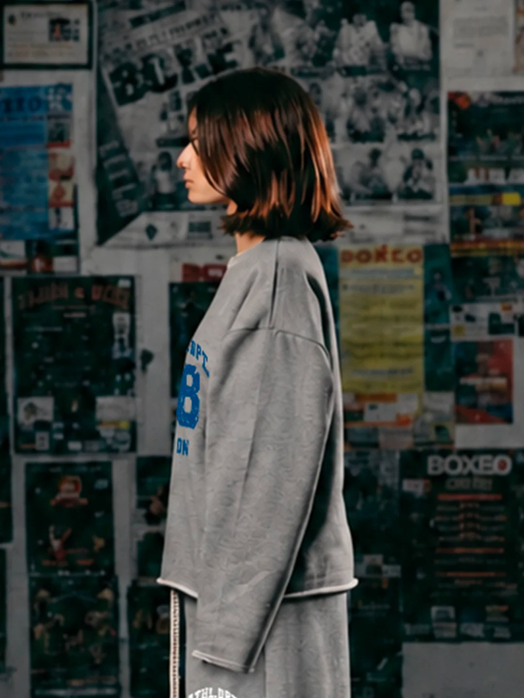 A woman wearing a grey oversized sweatshirt with blue and white lettering on the chest. She is standing in front of a wall covered in vintage boxing posters and flyers.