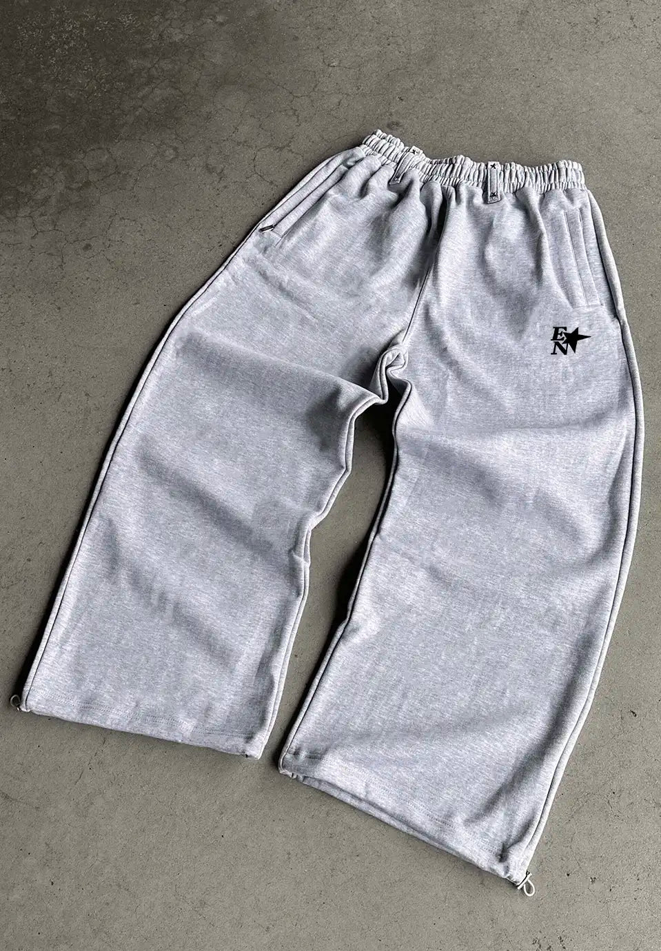 Light grey heather sweatpants laid flat on a grey concrete surface, featuring an elastic waistband, side pockets, and a small black embroidered star logo on the upper left leg.