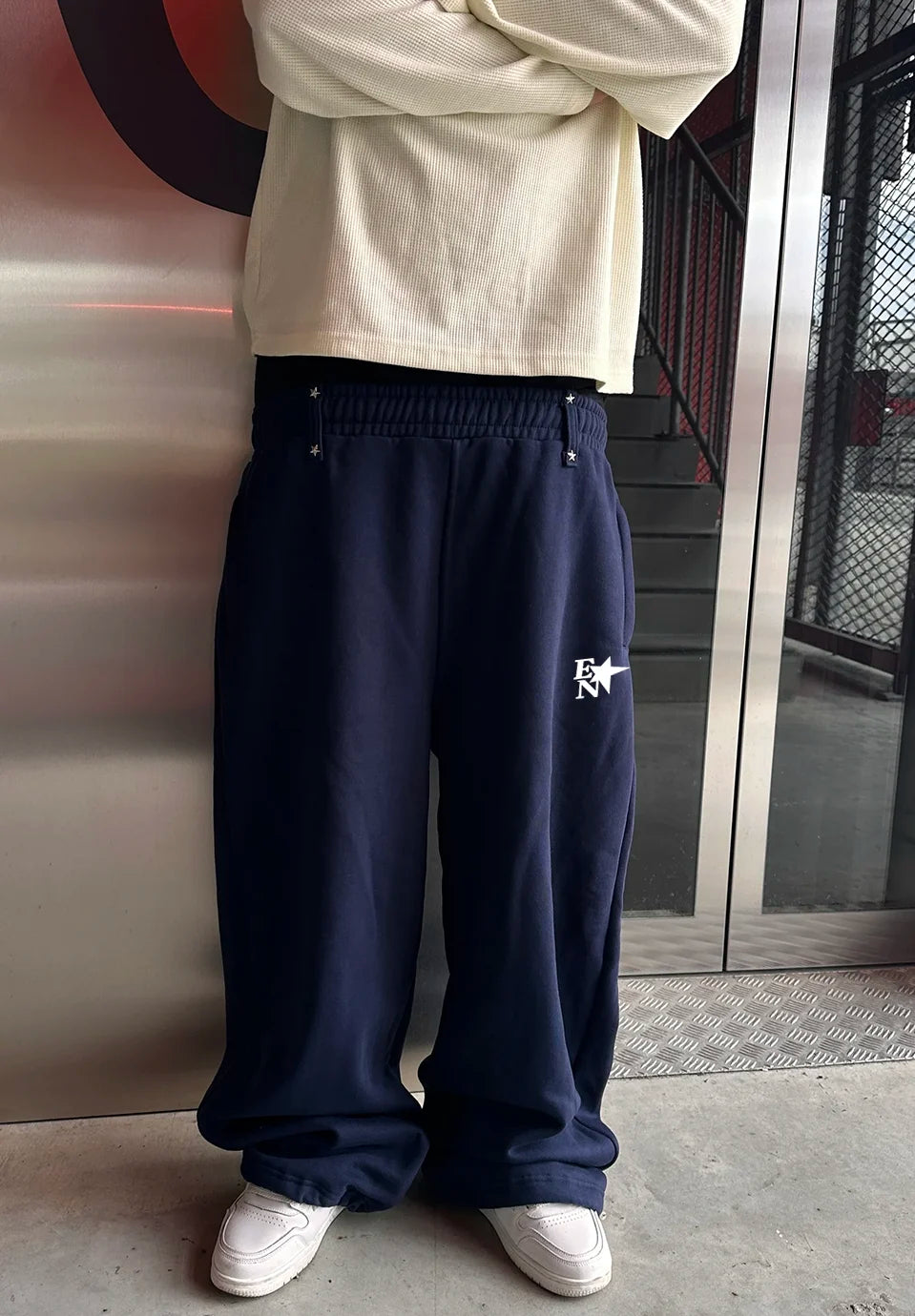 A person wearing loose navy sweatpants with a small white graphic logo on the left thigh, paired with a cream long-sleeve shirt and white sneakers, standing indoors near a glass door and metal wall.