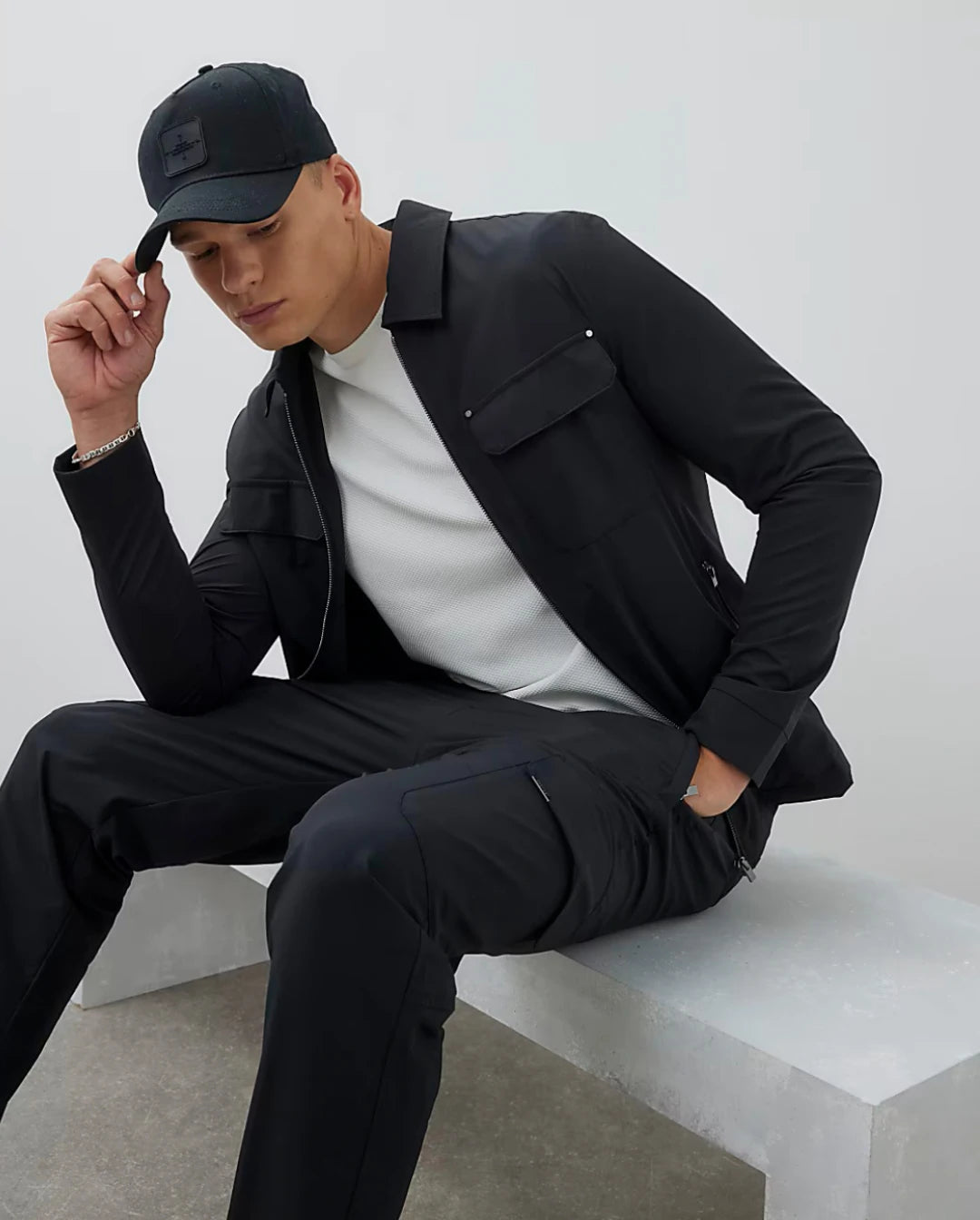 Man wearing a black jacket, white T shirt, and black pants sitting on a white surface.