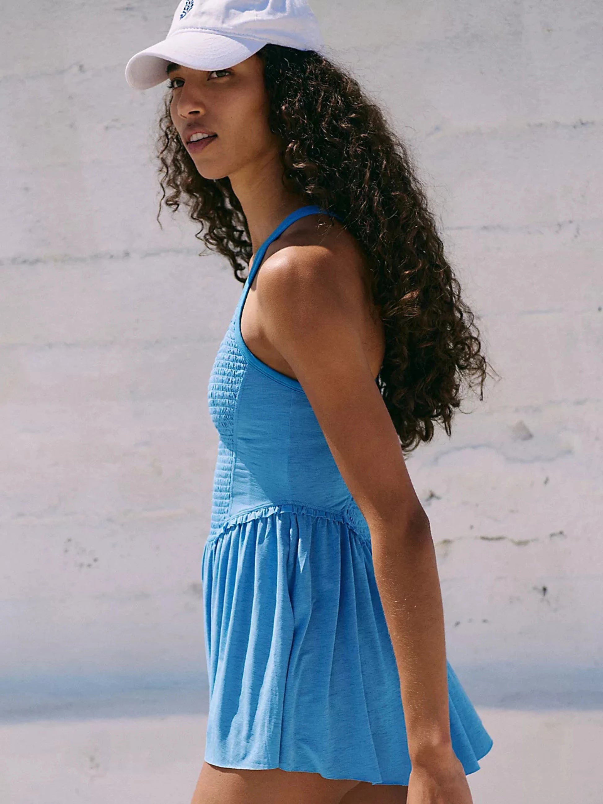 Woman wearing a blue dress and white cap against a light gray wall.