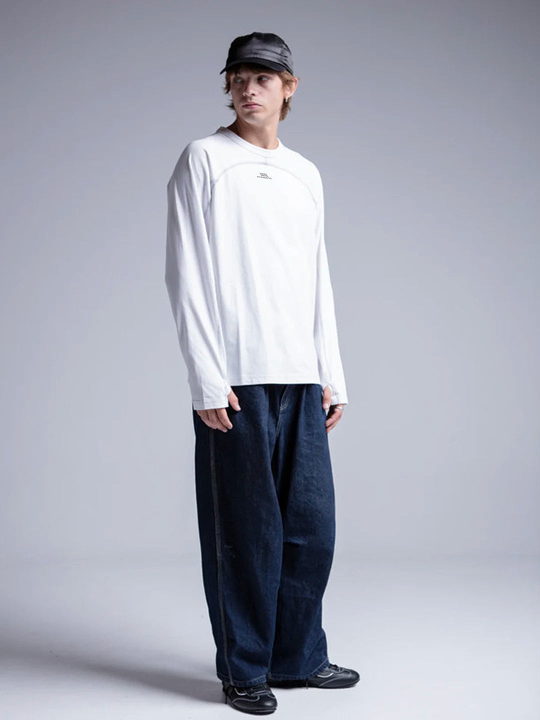 A model wearing a white long-sleeve crewneck shirt with a small chest logo, loose dark blue jeans, black shoes, and a black cap 