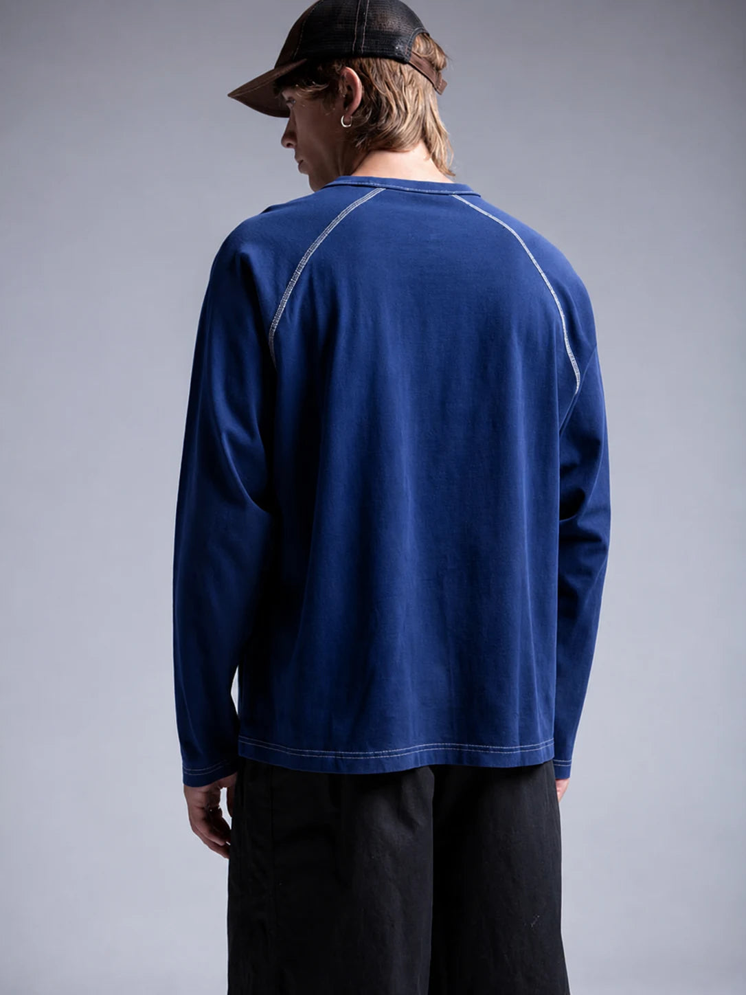 A man wearing a dark blue long-sleeve t-shirt with contrasting white stitching, black pants, and a black cap