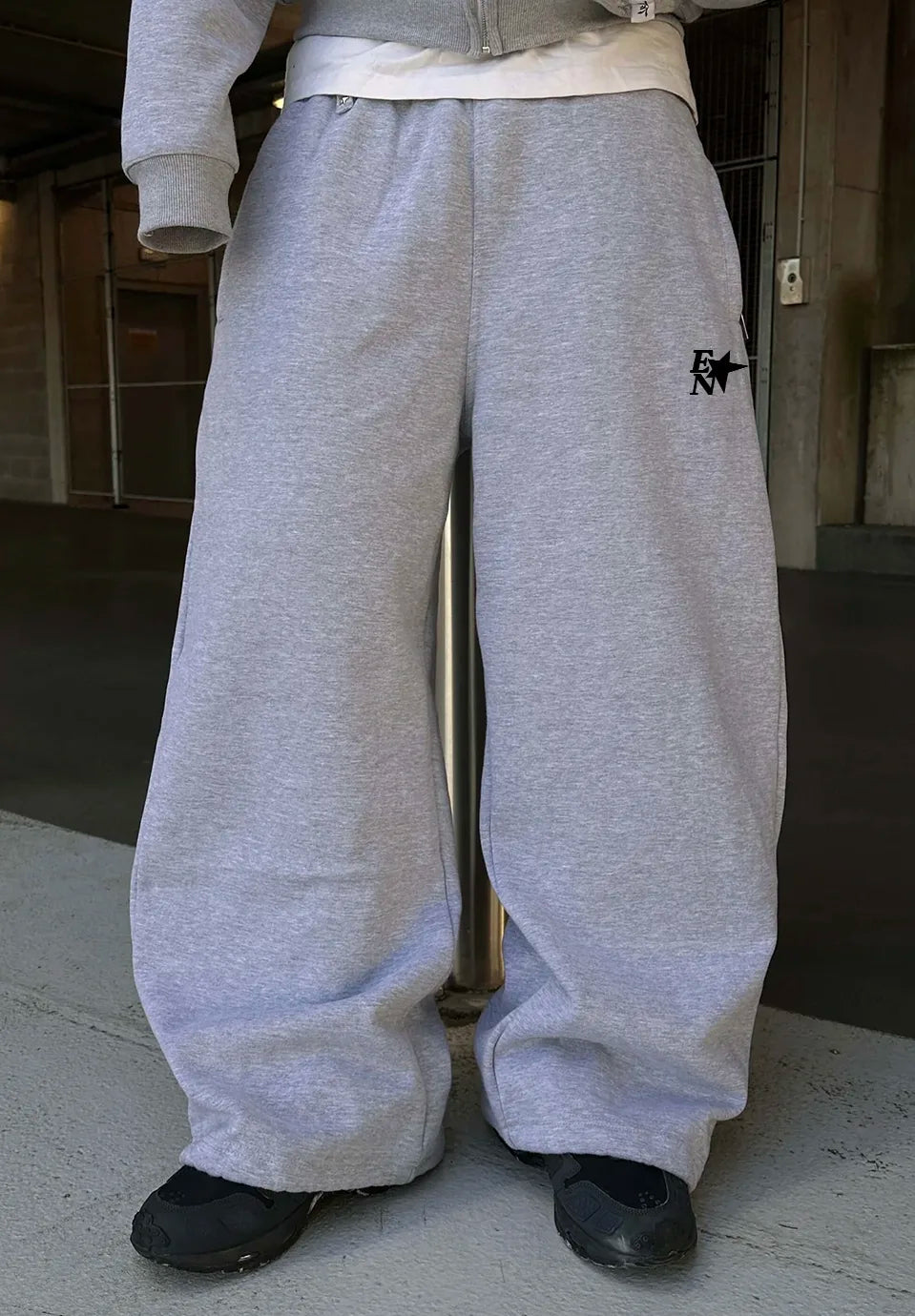 Grey wide-leg sweatpants with a white embroidered logo on the left thigh, worn by a model in an urban setting.