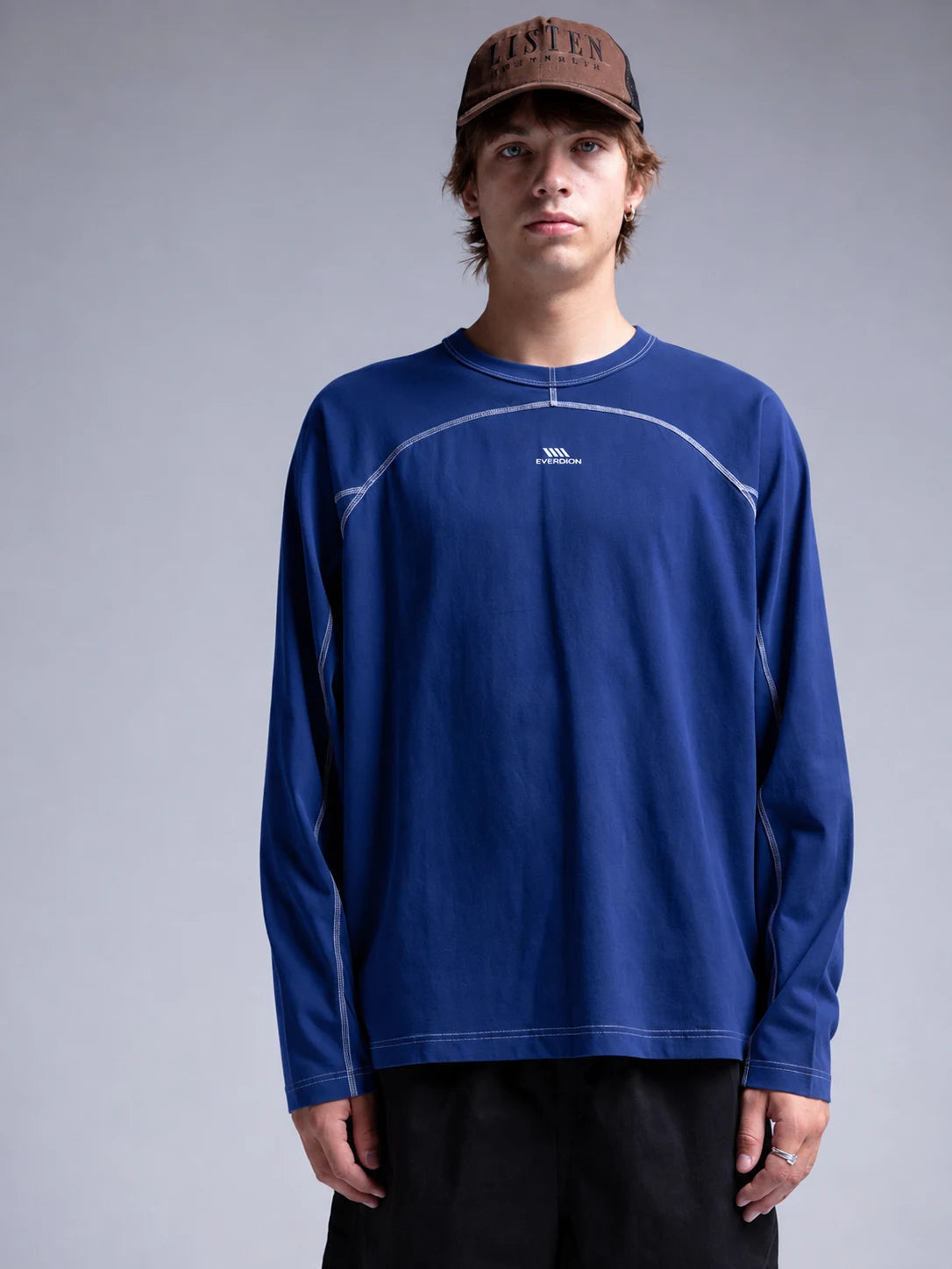 A model wearing a royal blue long-sleeve athletic t-shirt with white contrast stitching and a small white logo on the chest, paired with black pants and a brown cap