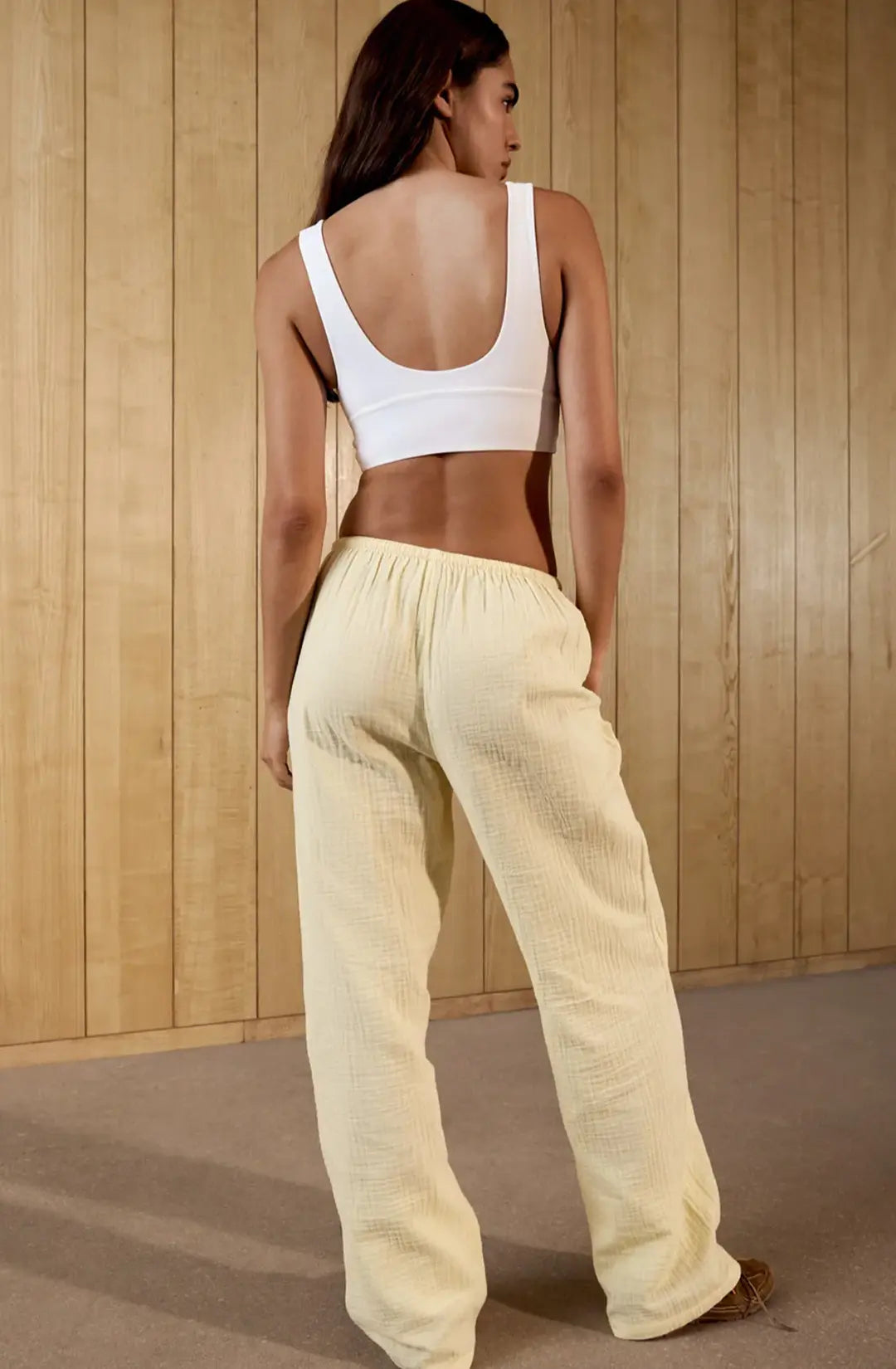 Woman wearing a white crop top and light-colored pants against a wooden wall.